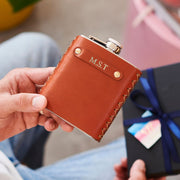 Person holding a brown leather flask with 'M.S.T.' initials, next to a gift box.
