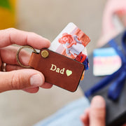 Brown keychain with 'Dad' and heart engraving held by a hand, with blurred gift box in the background.