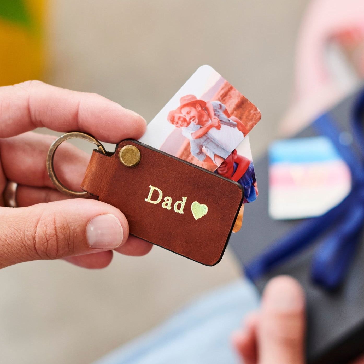 Brown keychain with 'Dad' and heart engraving held by a hand, with blurred gift box in the background.