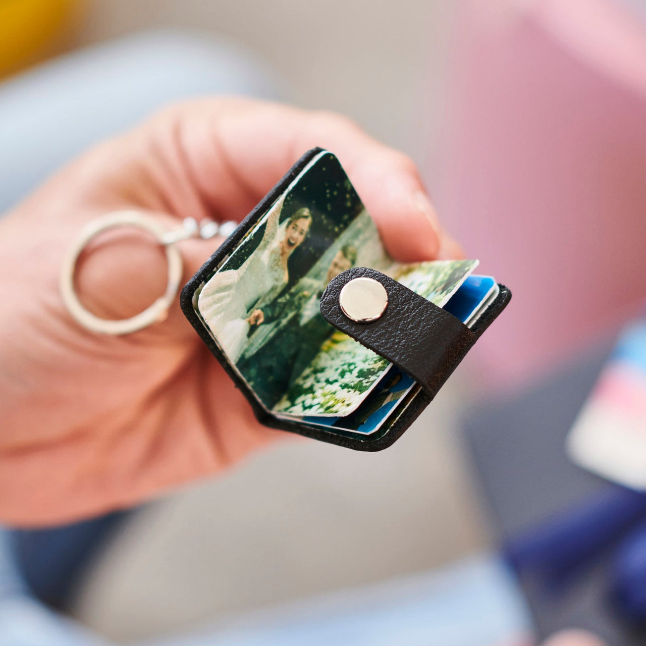 Hand holding a small leather book filled with photos with keychain attachment, blurred gift box in background