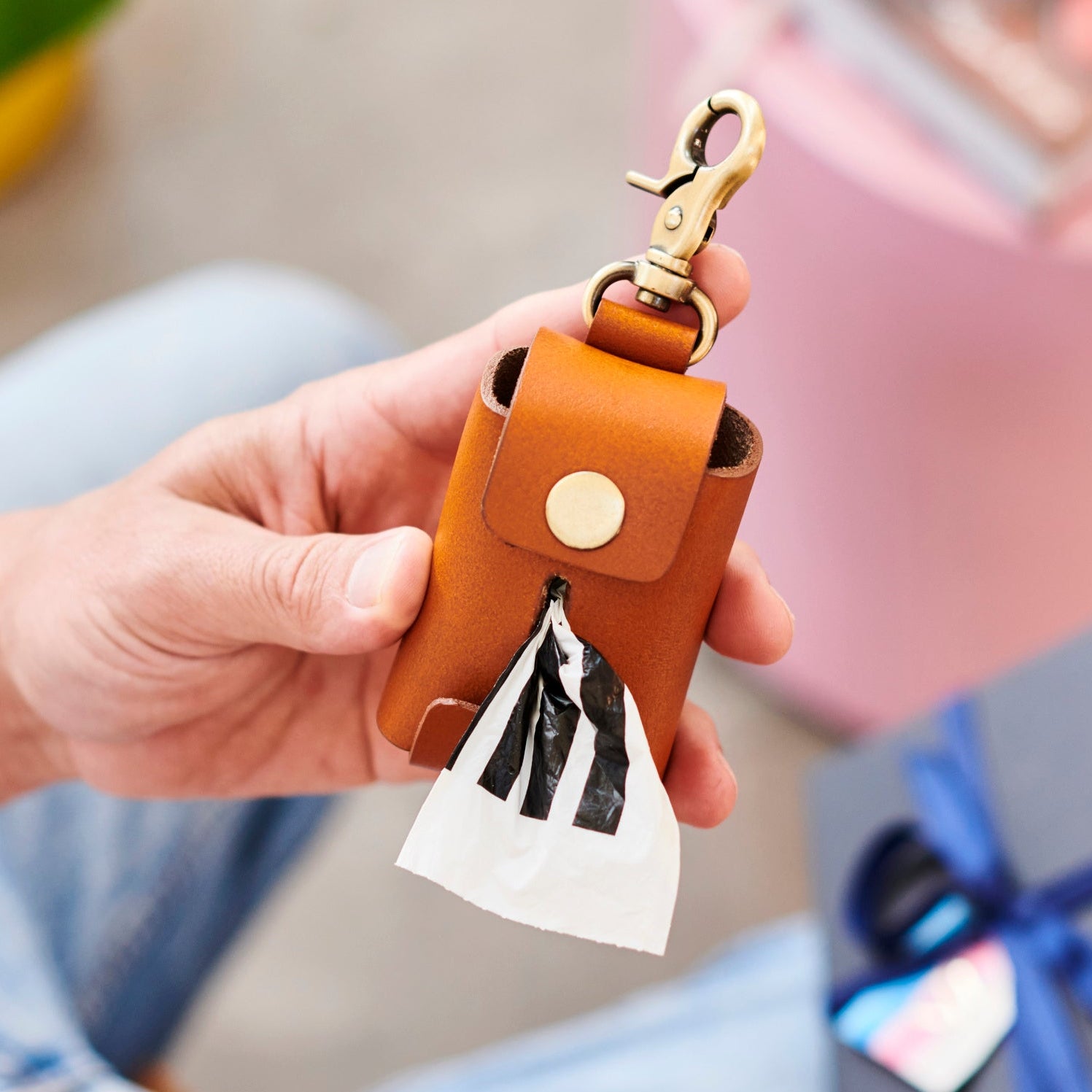 Person holding a tan leather holder for dog poo bags with metal clip. Ribbon tied gift Box blurred in the background. 